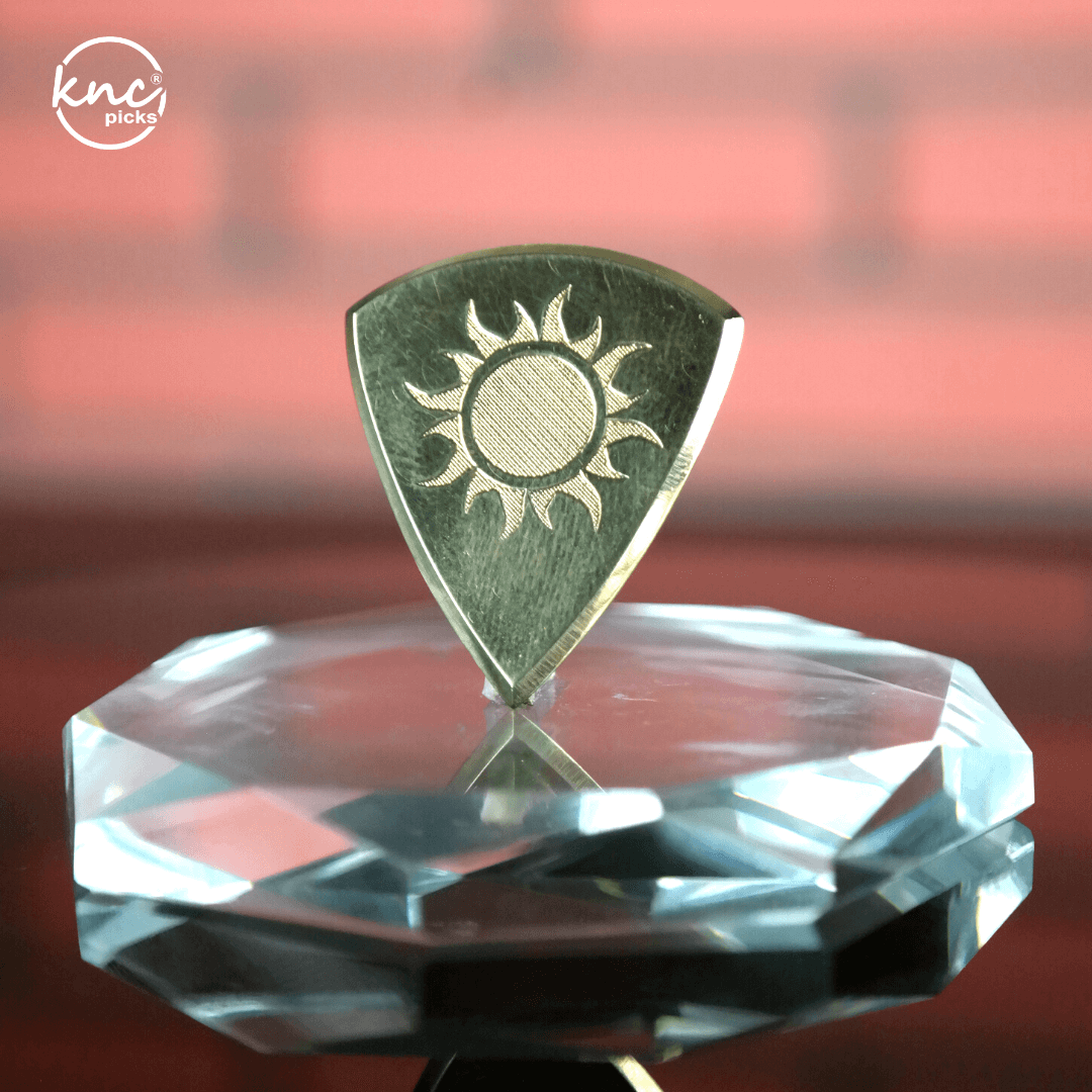 A Knc Picks Premium Brass Pick from the Summit Series, showing its polished, golden metallic finish and engraved logo.