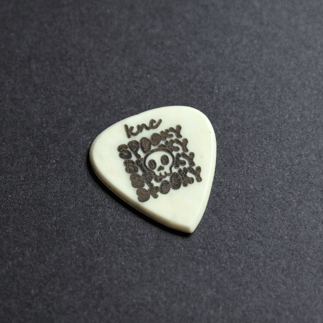 A Knc Picks handmade bone guitar pick featuring a spooky Halloween ghost design, showing its 2.0mm thickness.