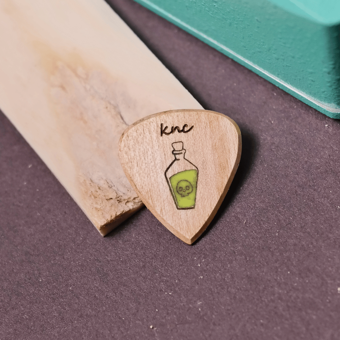 Close-up macro detail of the 'Poison Potion' bottle engraving on the natural maple wood guitar pick.
